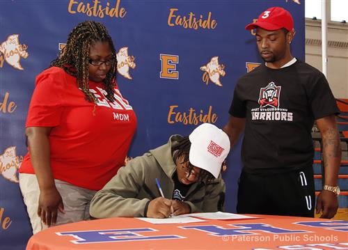Two people, male and female, watch as a young man wearing a baseball cap embroidered with "ESU", signs a paper while sitting.
